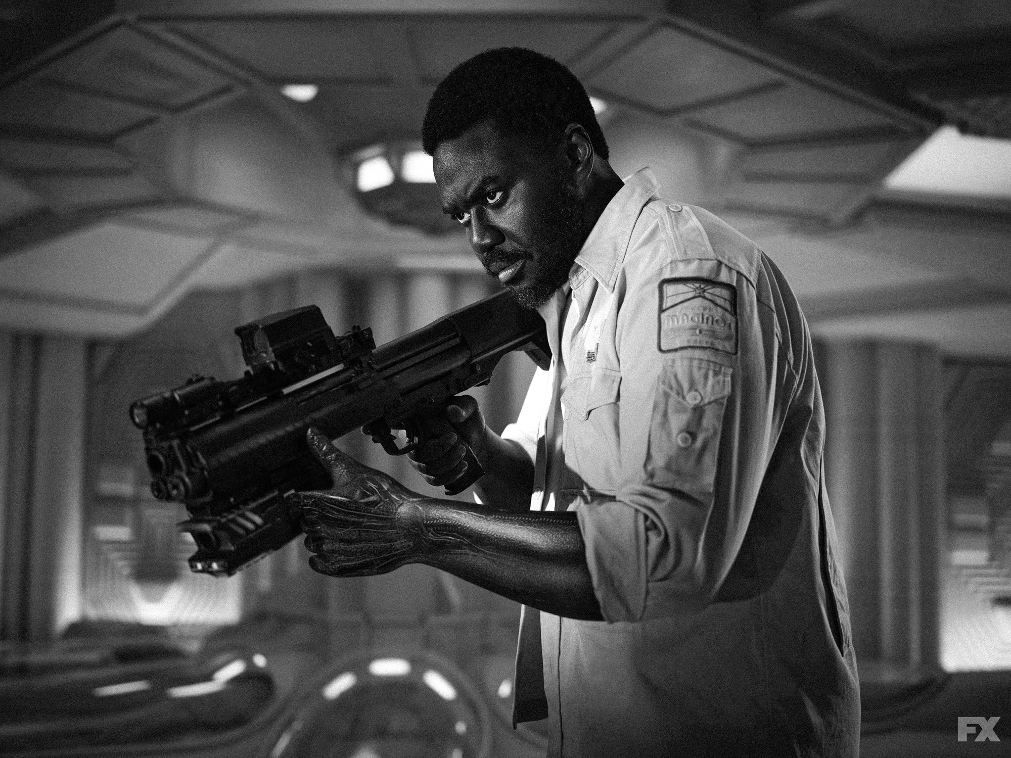 Mr. Morrow stands in a ship’s hypersleep chamber holding a gun and looking intently off camera.